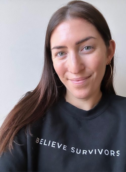 White woman with long brown hair, green eyes, gold hoop earrings, and a black sweatshirt that reads "believe survivors"