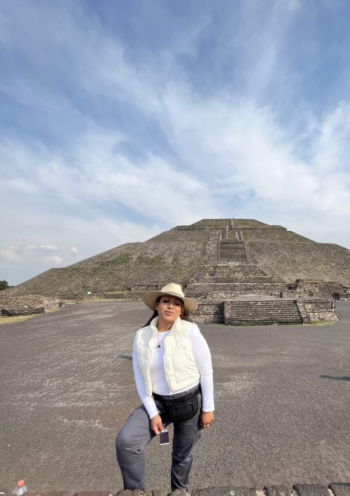Mexican woman with brown hair, a white hat, white shirt and vest, and grey pants standing in front of a Mayan pyramid.