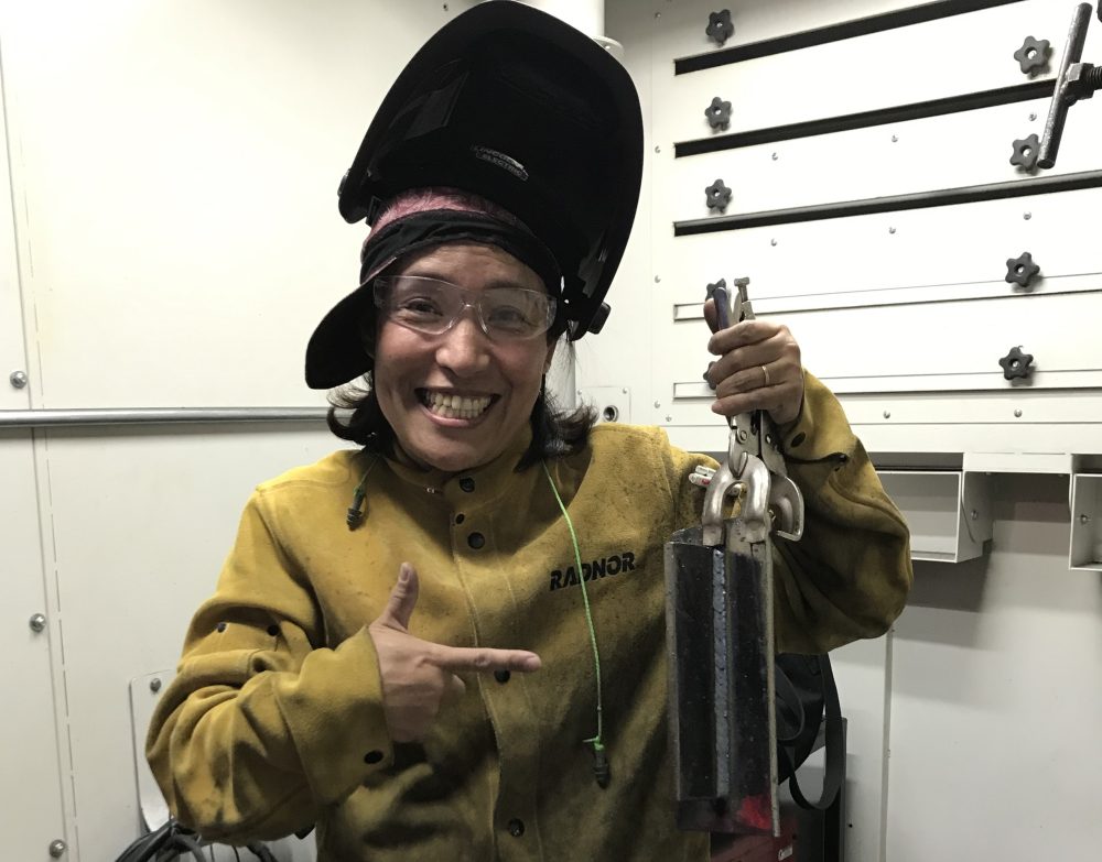 An IET Welding student wearing safety glasses, a welding helmet pushed back on their head, and a protective welding jacket smiles while holding up a metal object with clamps in a workshop. They are pointing proudly at the object, which appears to be a recently completed piece of welding work. Behind them, there is a stool and various workshop equipment, including tools and welding gear. The setting suggests an industrial or educational environment focused on metalworking or welding.