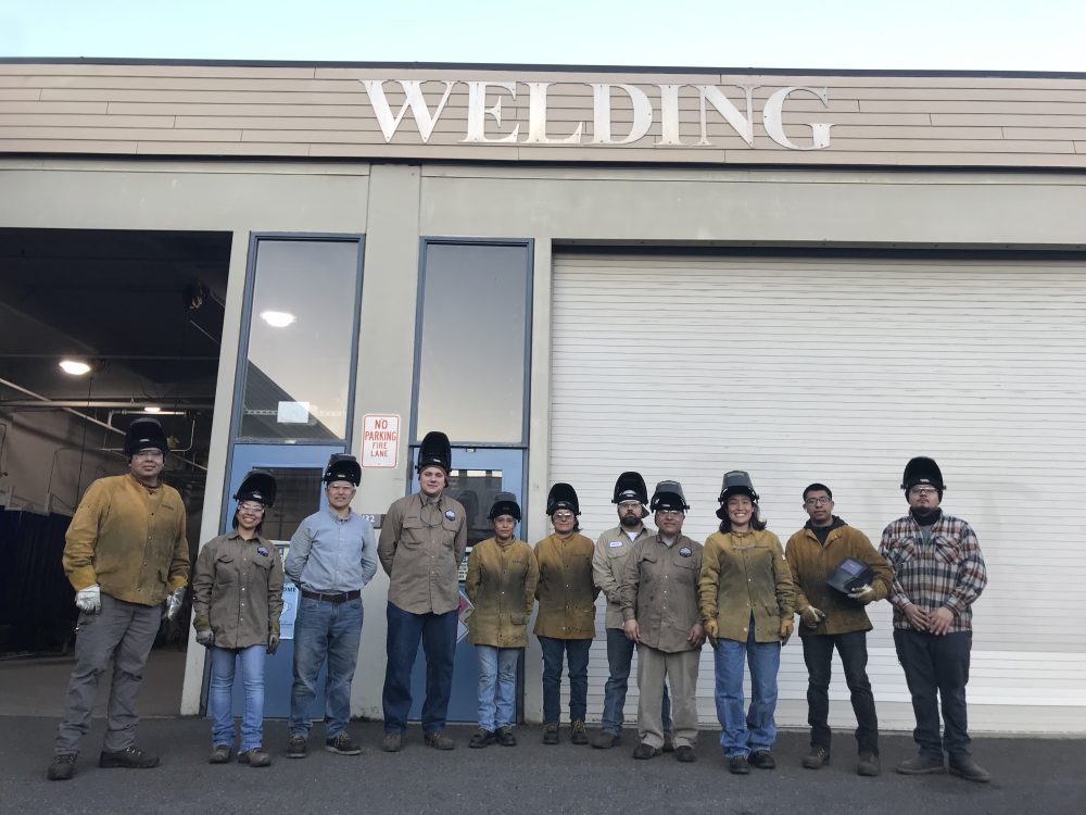 A group of IET Welding students and instructors wearing welding gear are standing outside the PCC Rock Creek Welding building with a sign that reads "WELDING".