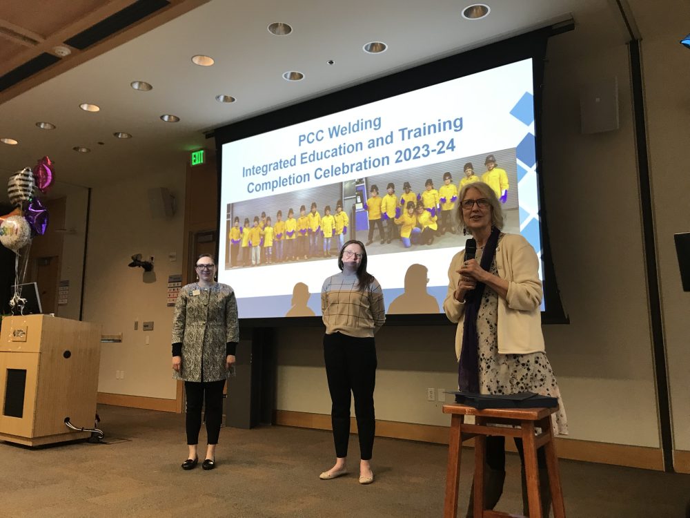 Three people are standing on a stage in front of a screen displaying "PCC Welding Integrated Education and Training Completion Celebration 2023-24." Balloons and a podium are seen in the background.