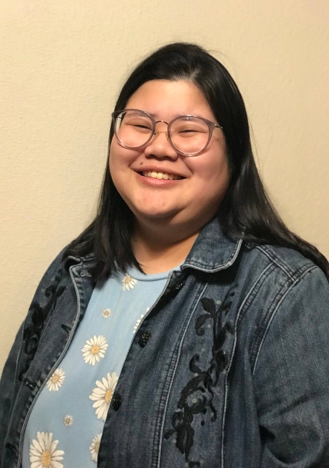 A half body profile image of a young Asian woman wearing glasses, Asher, stands in front of a blank backdrop, smiling at the camera. She wears a light blue blouse with white daisies of different sizes all over the blouse and a dark blue jean jacket with black plant embellishments on the chest.