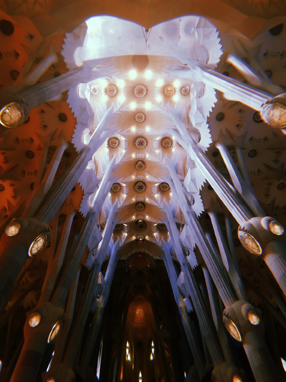 Inside of La Segrada Familia cathedral in Barcelona, Spain. The photo is taken inside the cathedral looking up at the roof. The room is largely dark on the sides but light shines through the roof.