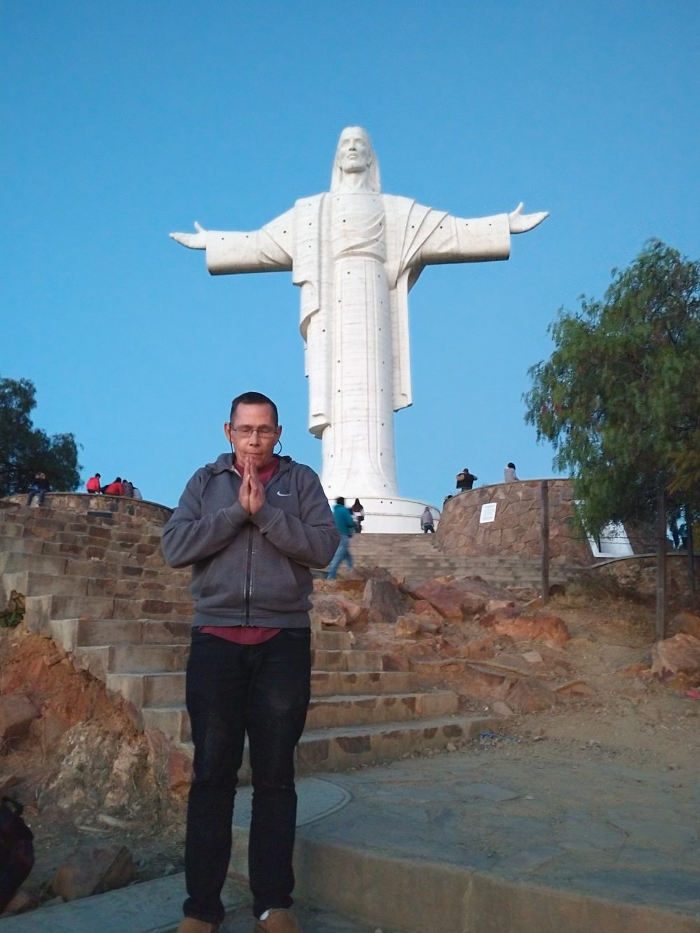 A male student wearing dark blue jeans, a red shirt, a gray Nike sweatshirt, brown shoes, and glasses with a string is holding his hands in prayer in front of the large, white Cristo de la Concordia statue of Jesus Christ in Cochabamba, Bolivia.