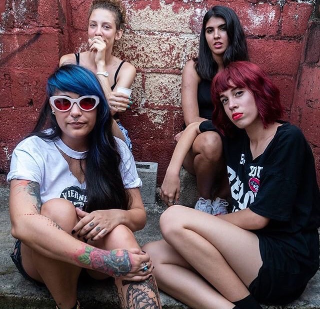 Four women sitting in a square pattern on the sidewalk in front of a red concrete brick wall with paint scratched off in the middle of the wall. The woman in the left foreground has a tattoo of a ship on her left leg and a tattoo of a rose on her right arm. She is wearing a white T-shirt that says Viernes and white-rimmed red sunglasses, and has dyed blue hair. In the right foreground, a woman with dyed red hair is wearing a black t-shirt and sitting sideways. In the left background, a woman with curly brown hair has her hand cupped over her mouth and is holding a white paper cup. In the right background, a woman with black hair and brown eyes is looking at the camera and is seen wearing a black shirt and white tennis shoes.