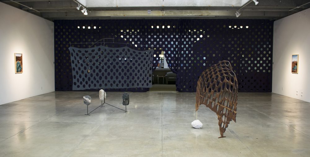 A sculpture on the left has 3 stones at its base and a grey screen with holes in it; a sculpture on the right has a wooden screen with holes in it standing on end with a stone as a base on one side. 2 landscape photos hand on either side; behind is a floor to ceiling, wall to wall dark curtain with a grid of large holes in it.