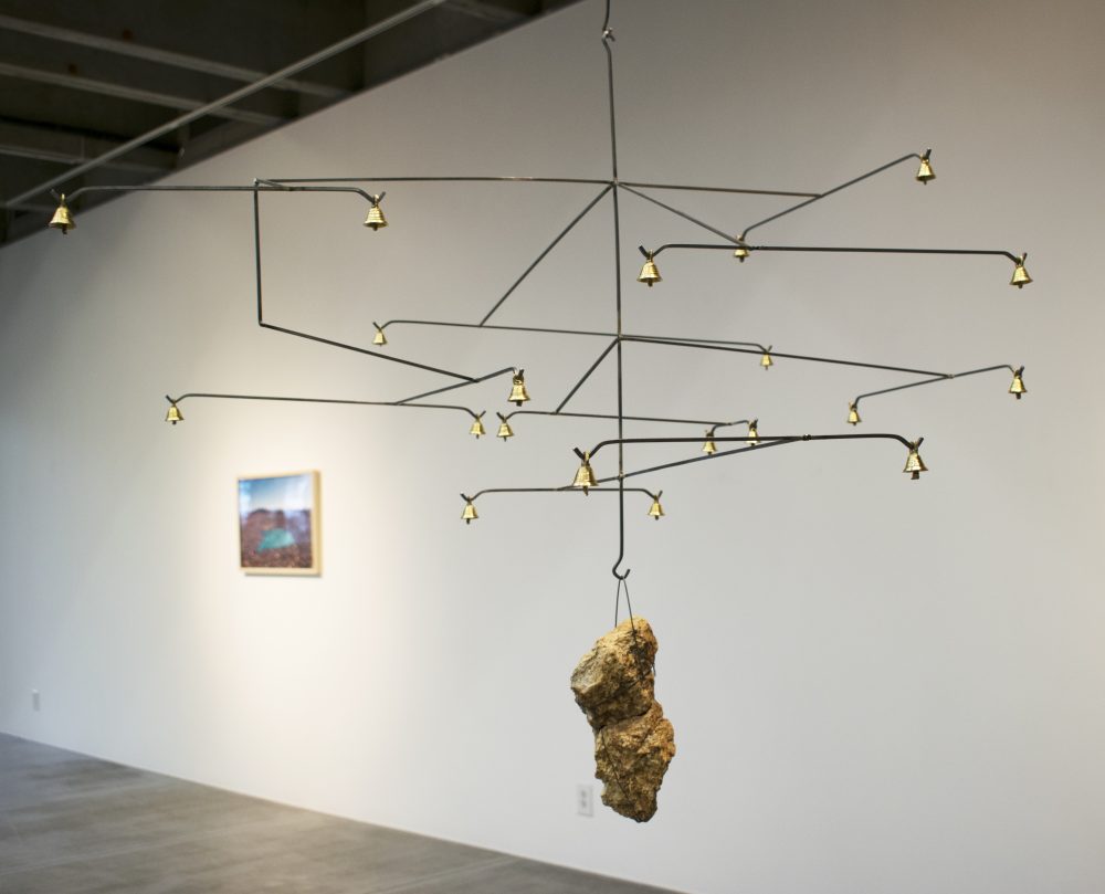 A mobile sculpture made of horizontal wires with bells on either end, and a center wire that holds a rock at the bottom, is in the foreground of the picture. Behind it is a gallery wall with a landscape photo.