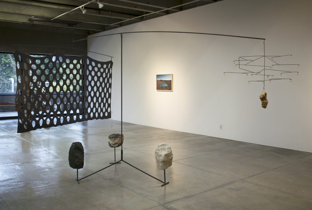 Large sculpture made with steel rods; 3 large rocks are at the base; it has a wide screen with large holes on the left, and a wire mobile with bells on the right. In the background is a wall with a landscape photo hanging on it.