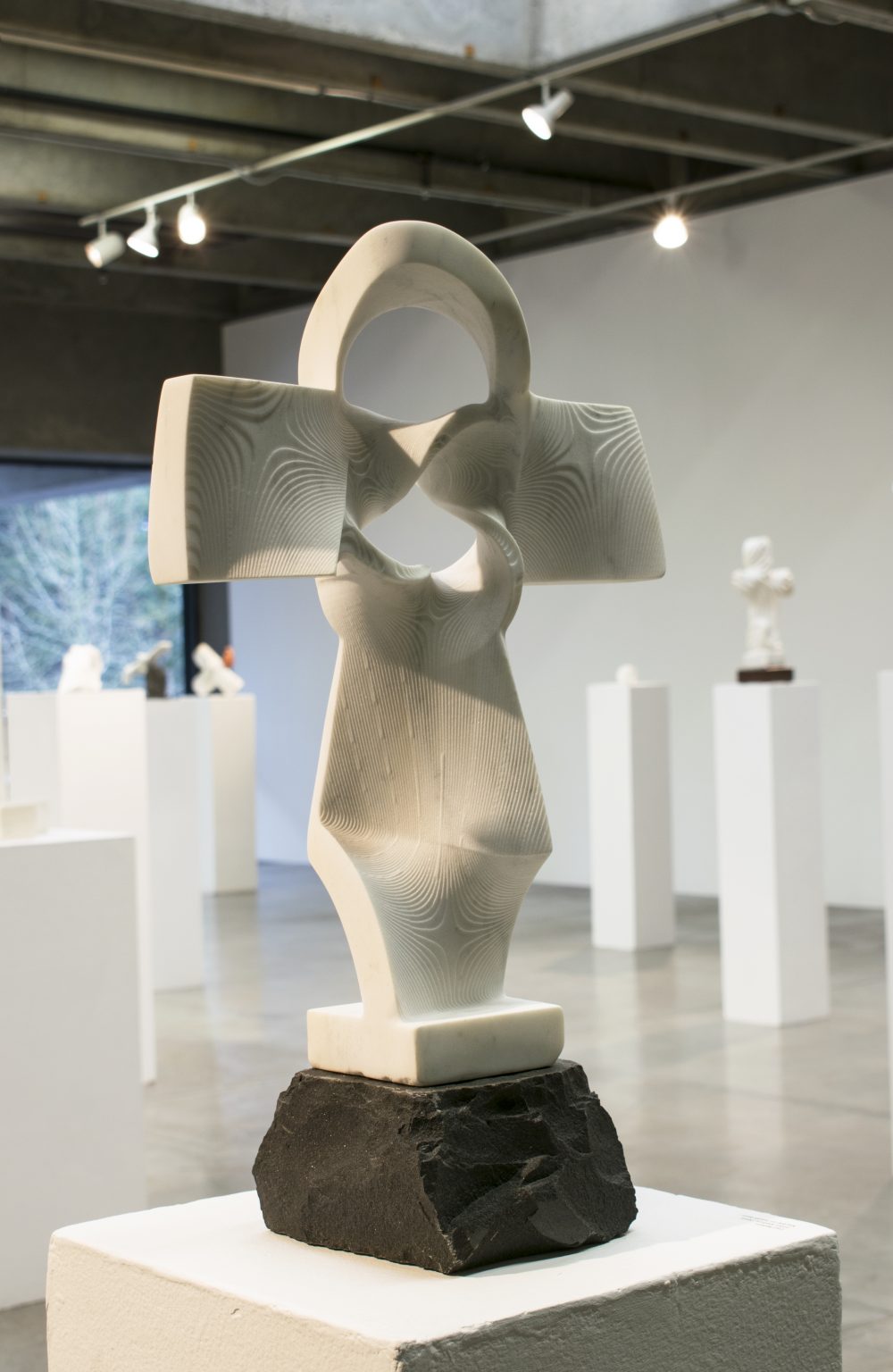 Marble sculpture in shape of a twisted cross, on a base of dark basalt, on pedestal, with other sculptures on pedestals in background.