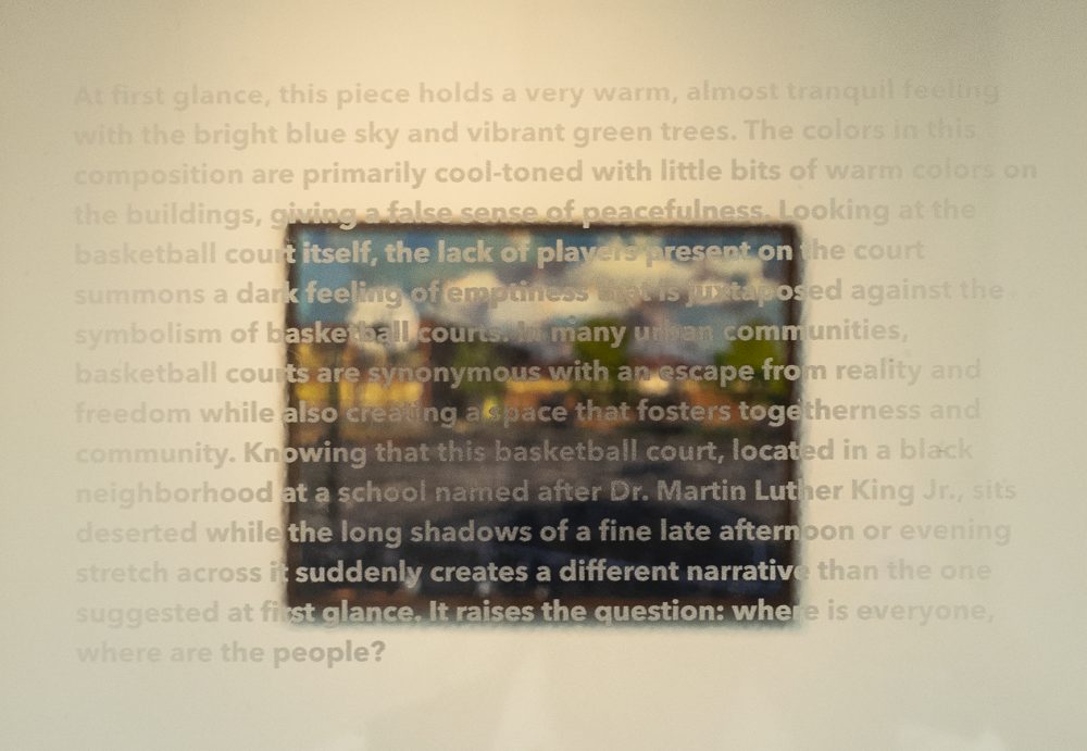 A representational painting of a schoolyard basketball court is hanging on a wall in the background. In the foreground is text describing the painting, printed on a clear acrylic panel which is placed directly in front of it.