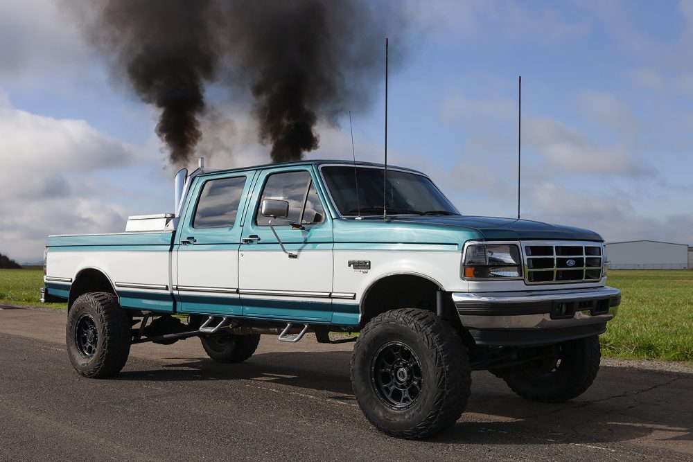 Photograph of a blue and white pickup truck driving on a road. The view is from the side of the truck, asphalt in the foreground, grass and partly cloudy sky in the background. Two dark black plumes of smoke rise from the exhaust pipes on the back of the cab of the truck.