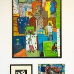 Three artworks hang on a wall. On top is a large multicolored, vertical, abstract painting depicting people in the street. On the bottom left is a small black and white print of three seated figures; on the bottom right a square painting of a woman sleeping in a bed with a child on either side of her. The painting is predominantly blue.