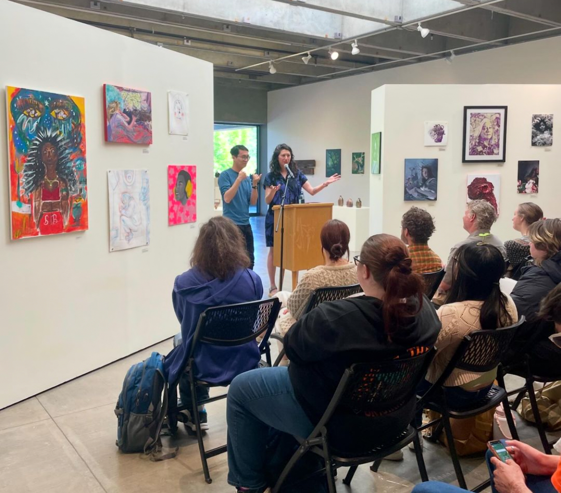 Two artists talk at a podium in an art gallery in front of an audience of students.