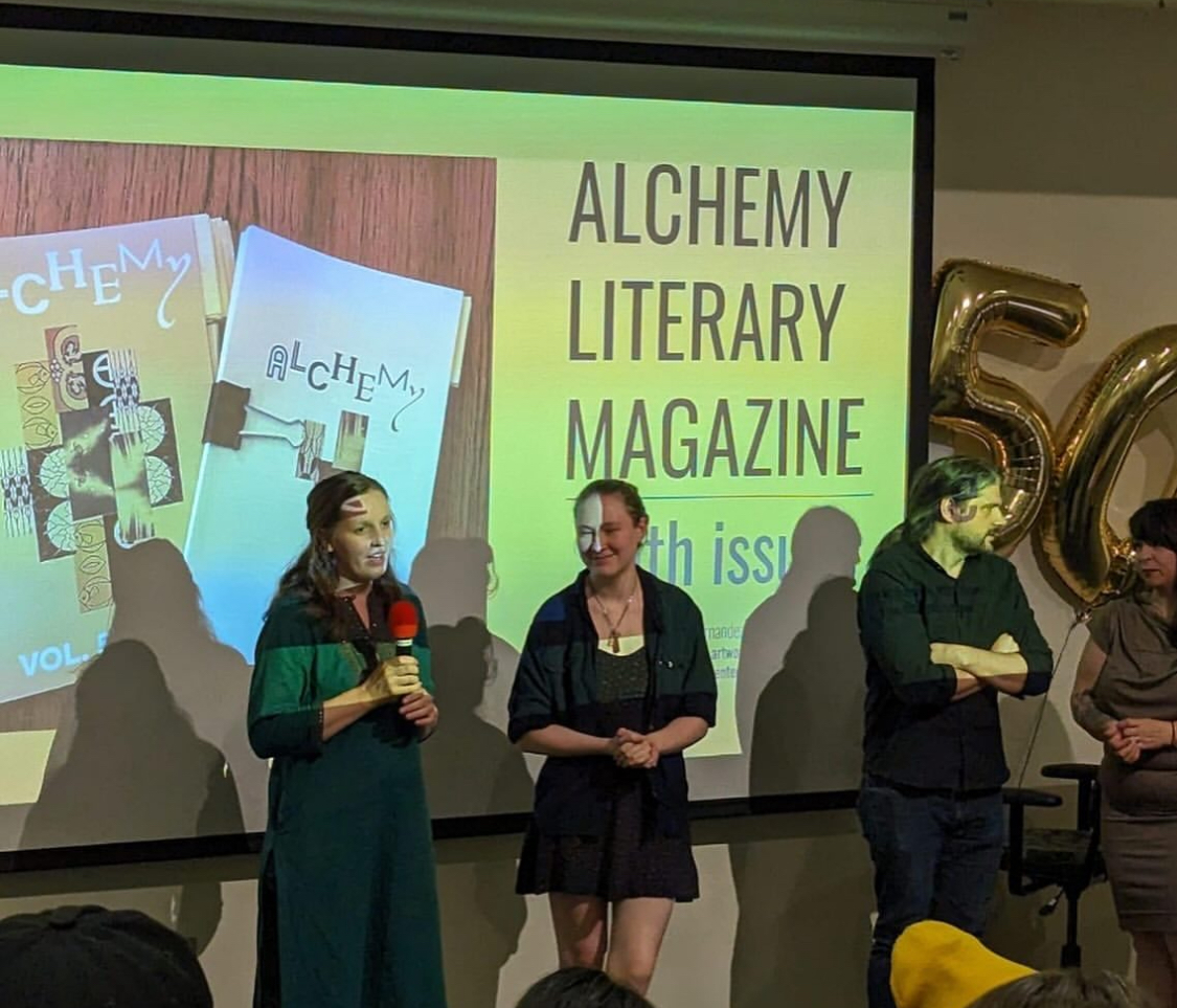 Two students stand on stage in front of a presentation. The presentation features an image of a student-made literary magazine.