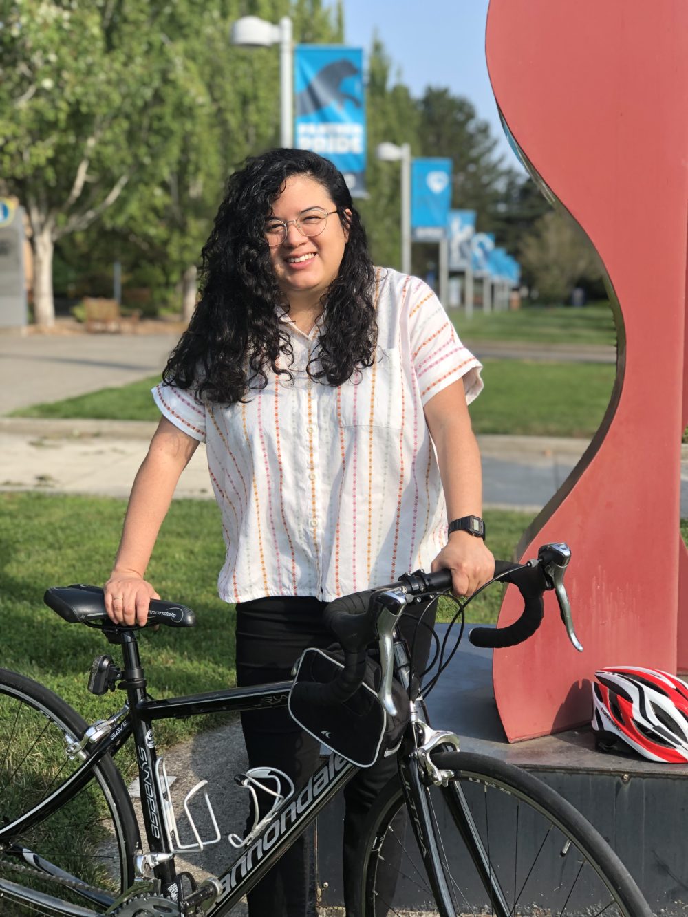 Andrea Reyes stands with her bicycle next to a red sculpture. Grass, trees and PCC signs are in the background.