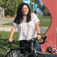 Andrea Reyes stands with her bicycle next to a red sculpture. Grass, trees and PCC signs are in the background.