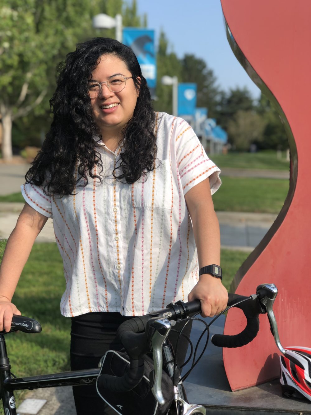 Andrea Reyes stands with her bicycle next to a red sculpture. Grass, trees and PCC signs are in the background.