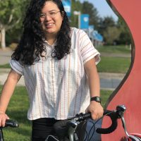 Andrea Reyes stands with her bicycle next to a red sculpture. Grass, trees and PCC signs are in the background.