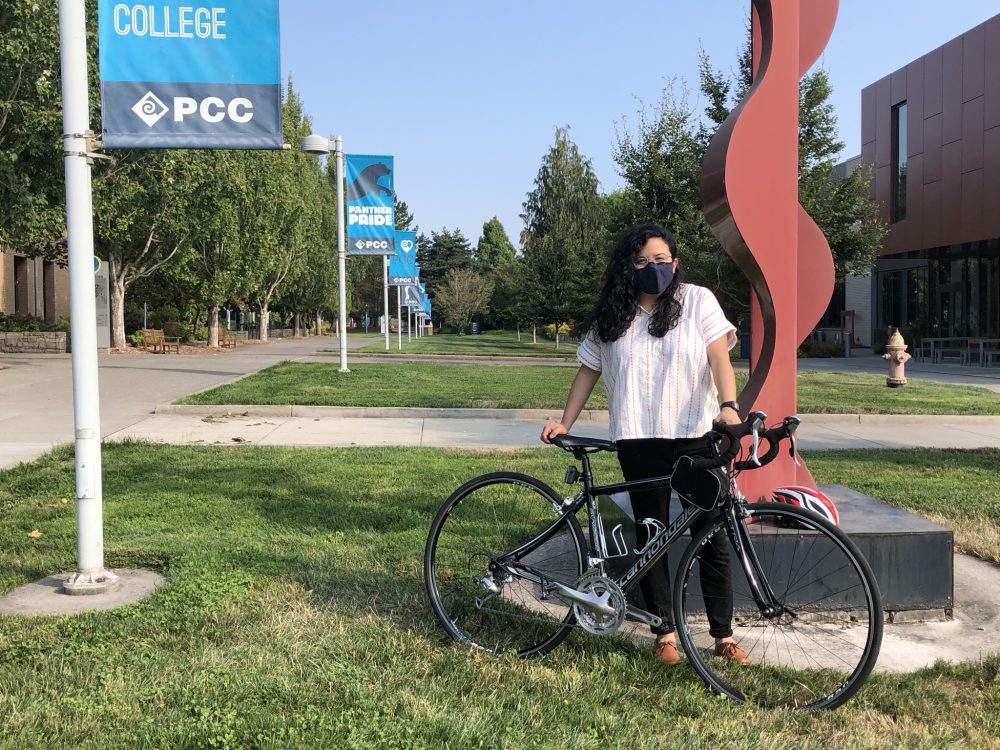 Andrea Reyes stands with her bicycle next to a red sculpture. Grass, trees and PCC signs are in the background.