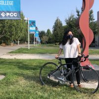 Andrea Reyes stands with her bicycle next to a red sculpture. Grass, trees and PCC signs are in the background.