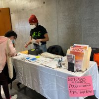 Table with resources and pamphlets. Person standing behind the table assisting another person looking at the information.
