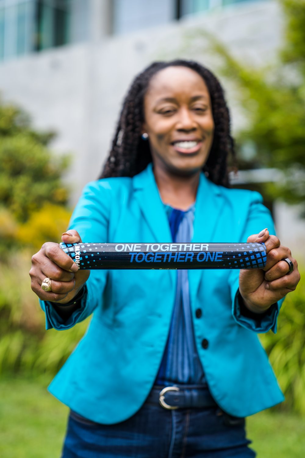 Dr. Adrien Bennings standing outside wearing a PCC turquoise blazer and holding a "One Together, Together One" prop up to the camera