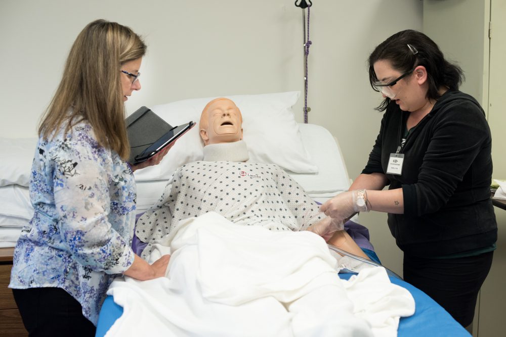 An instructor coaches a female student as she practices entry level CNA skills on healthcare mannequin in bed