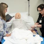 An instructor coaches a female student as she practices entry level CNA skills on healthcare mannequin in bed