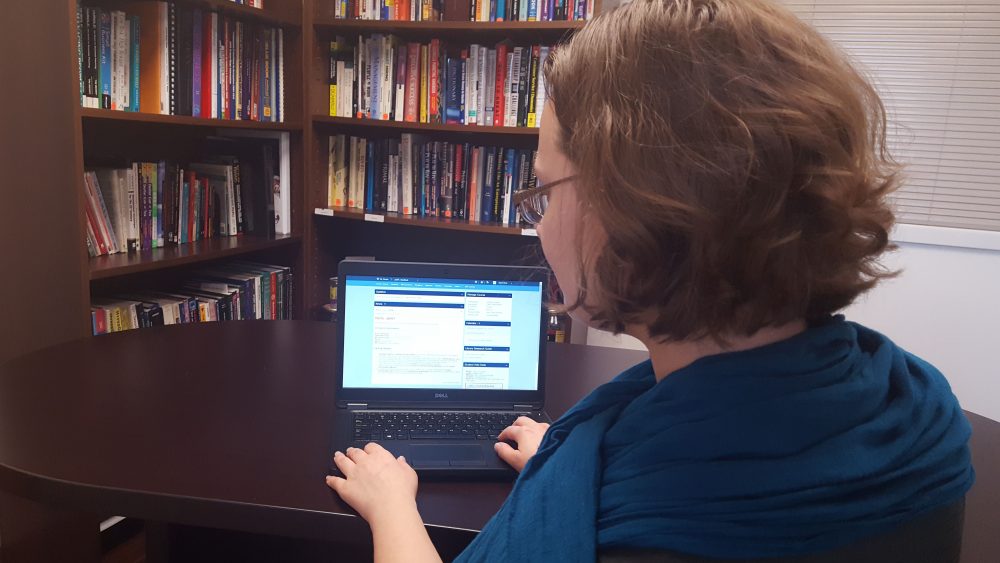 Woman with blue scarf sits at a desk in front of laptop with PCC online course on screen