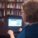 Woman with blue scarf sits at a desk in front of laptop with PCC online course on screen