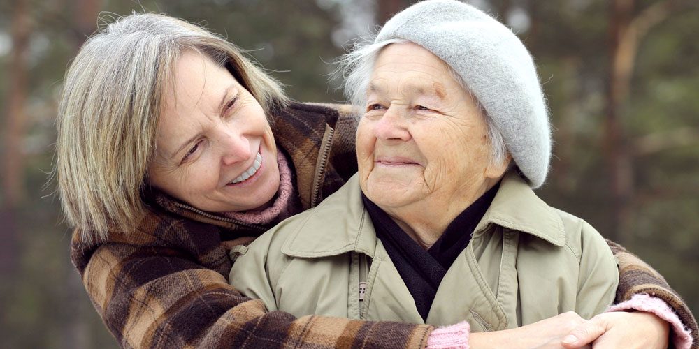 Aging adult mother looks up as she is hugged by her adult daughter