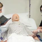 An instructor coaches a female student as she practices entry level CNA skills on healthcare mannequin in bed