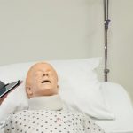 An instructor coaches a female student as she practices entry level CNA skills on healthcare mannequin in bed