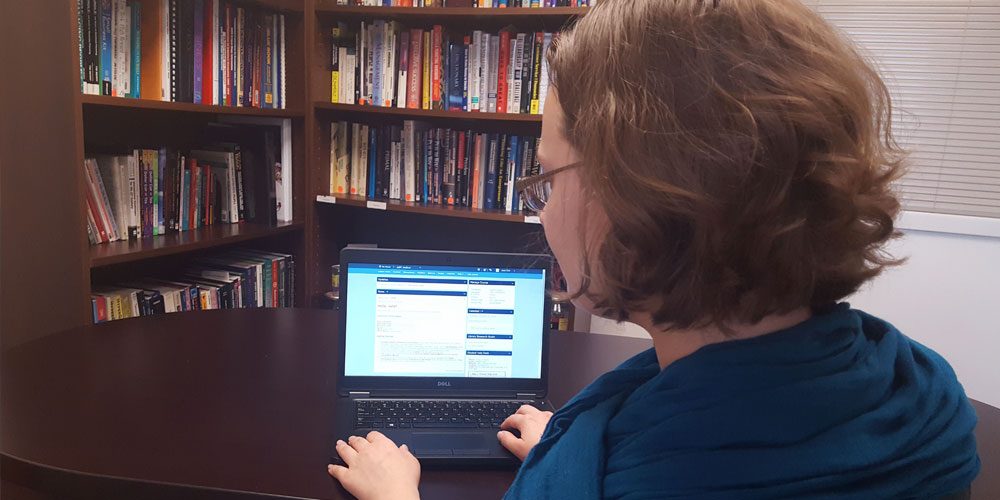 Woman with blue scarf sits at a desk in front of laptop with PCC online course on screen