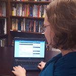 Woman with blue scarf sits at a desk in front of laptop with PCC online course on screen