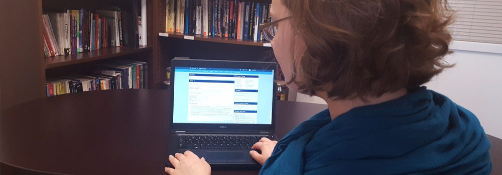 Woman with blue scarf sits at a desk in front of laptop with PCC online course on screen