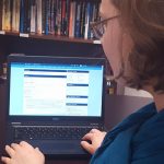 Woman with blue scarf sits at a desk in front of laptop with PCC online course on screen