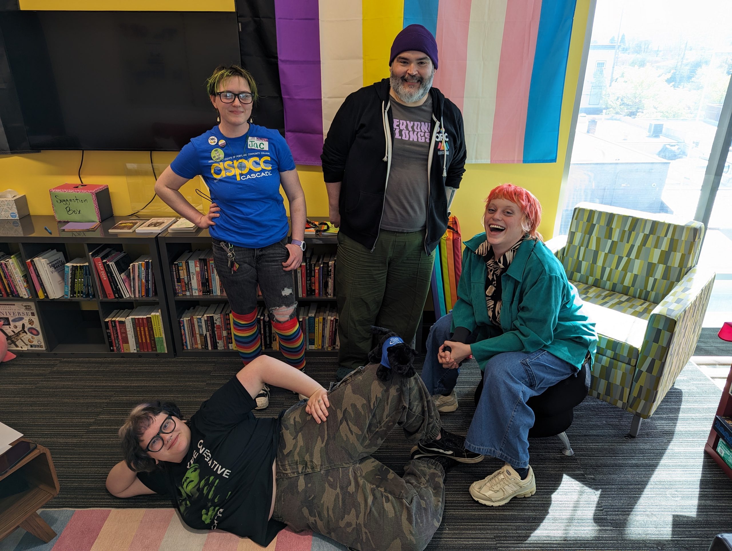 Four students, (two standing, one seated, and one lying on the floor) in front of a backdrop of rainbow flags