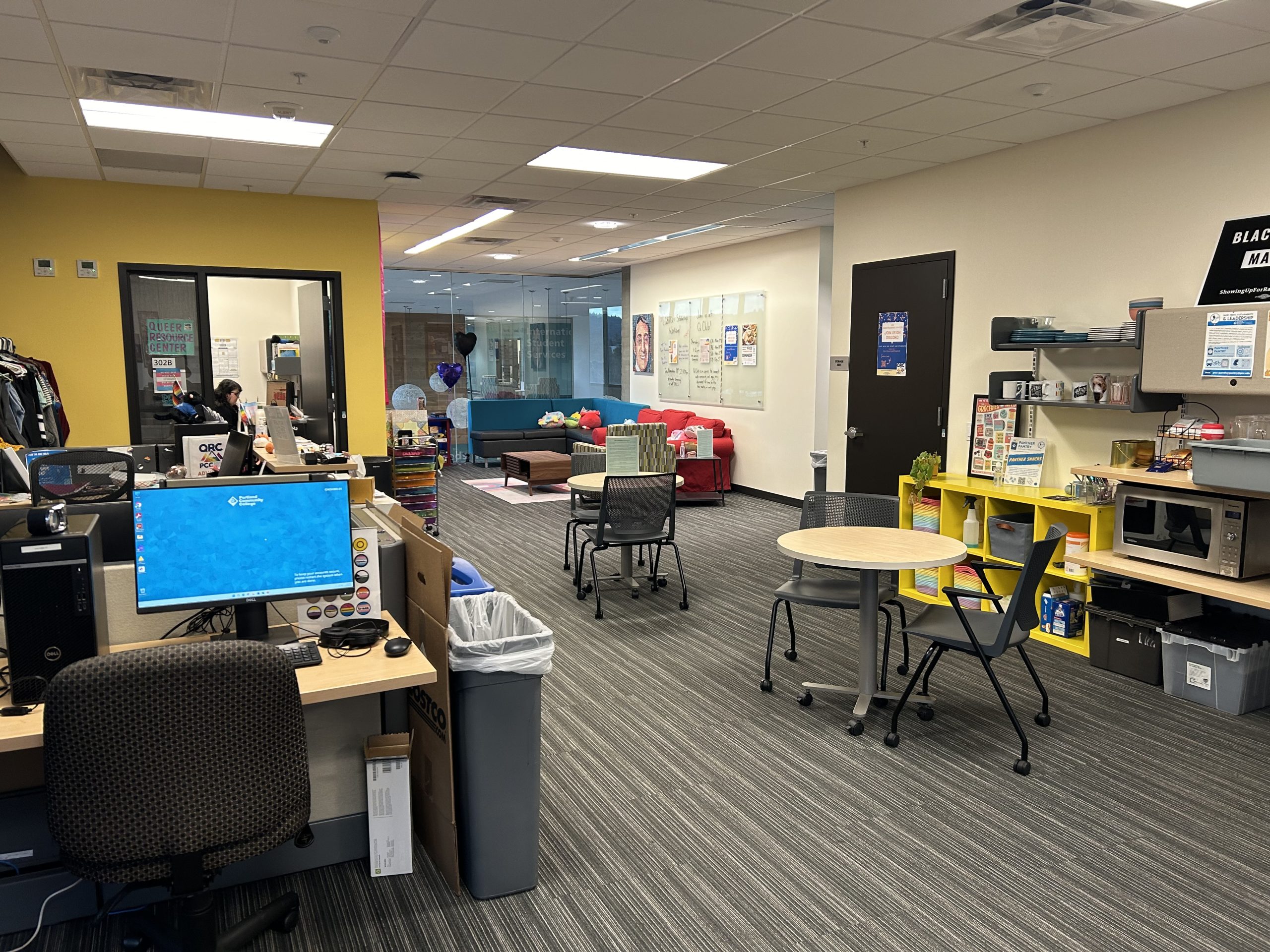 A room with one yellow wall, a computer desk in the foreground, table and chairs next to a kitchen area, couches and a glass wall in the background. 