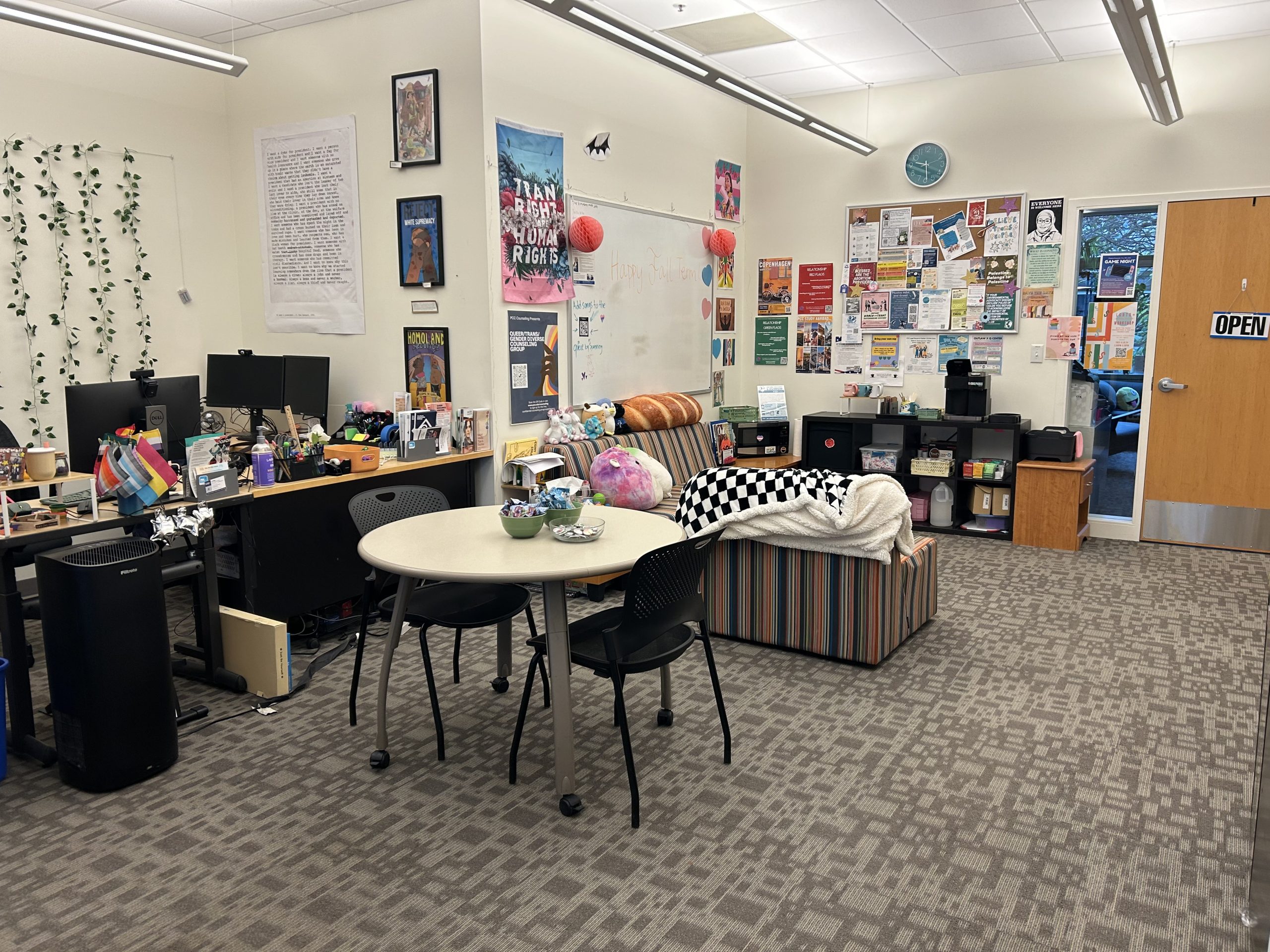 A view of a room with a desk, a table with chairs, and two couches. There are many posters on the wall. 