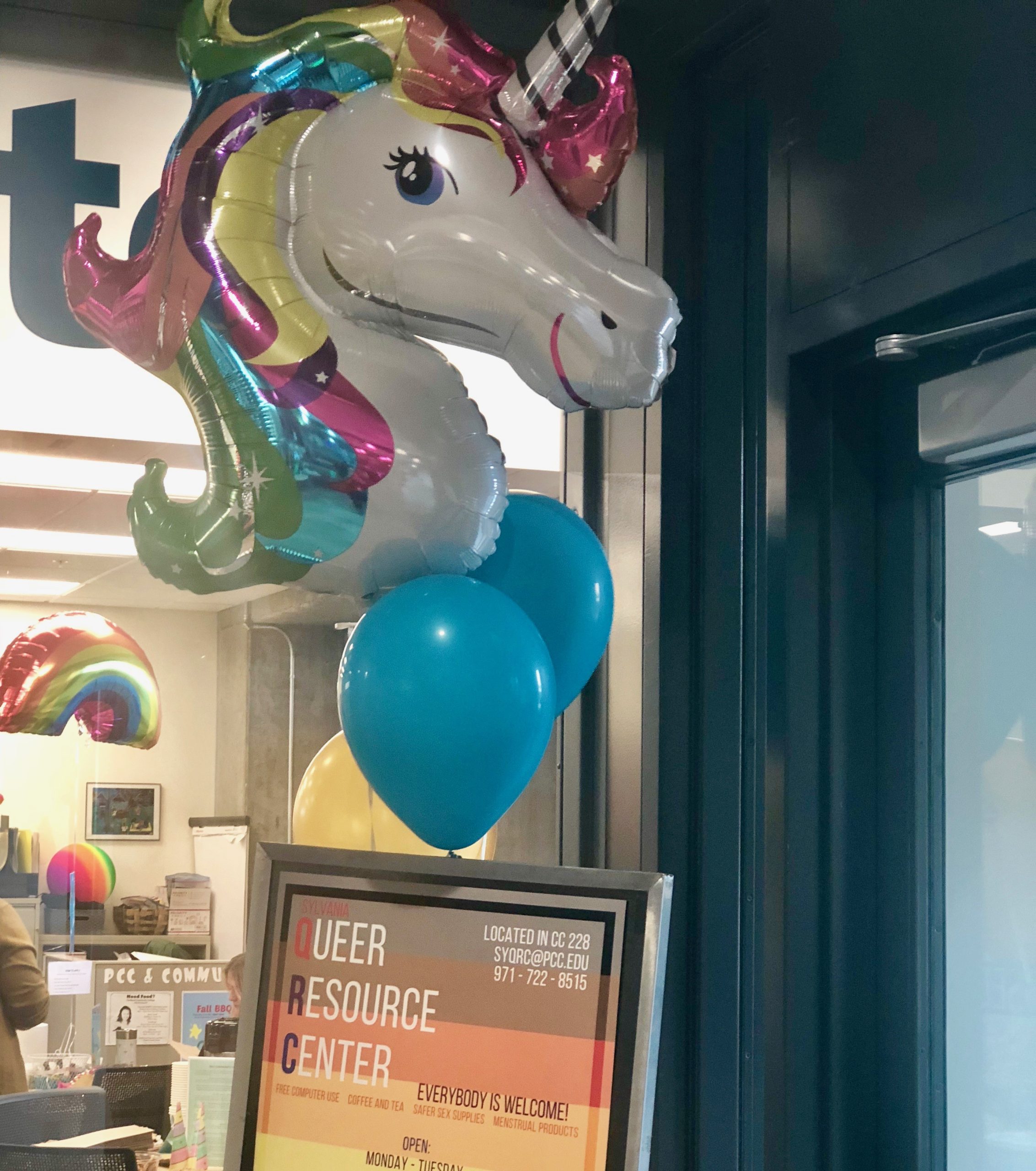 A large unicorn balloon with a rainbow main is attached to a sign for the Queer resource center. Behind the balloon is a window looking into the Sylvania QRC. 