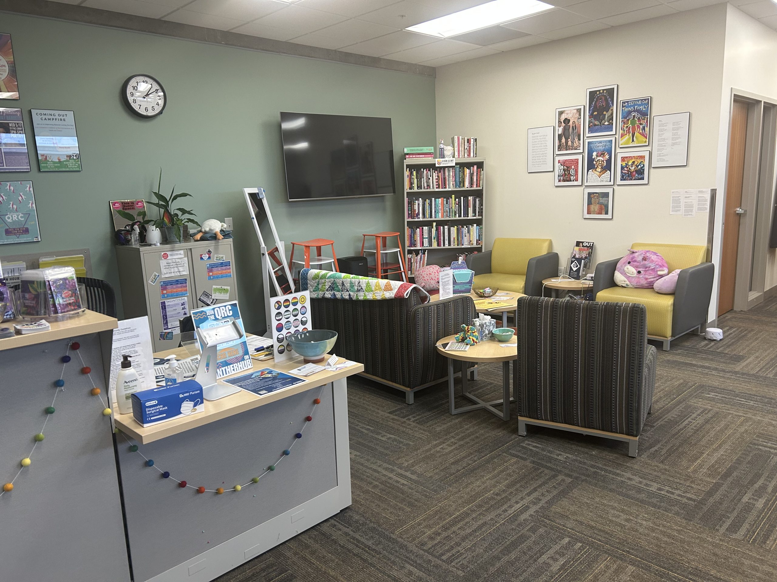 A photo of a colorful lounge space with couches, coffee tables, a bookcase and a TV Screen. In the foreground is a receptions area with flyers and posters on it. 